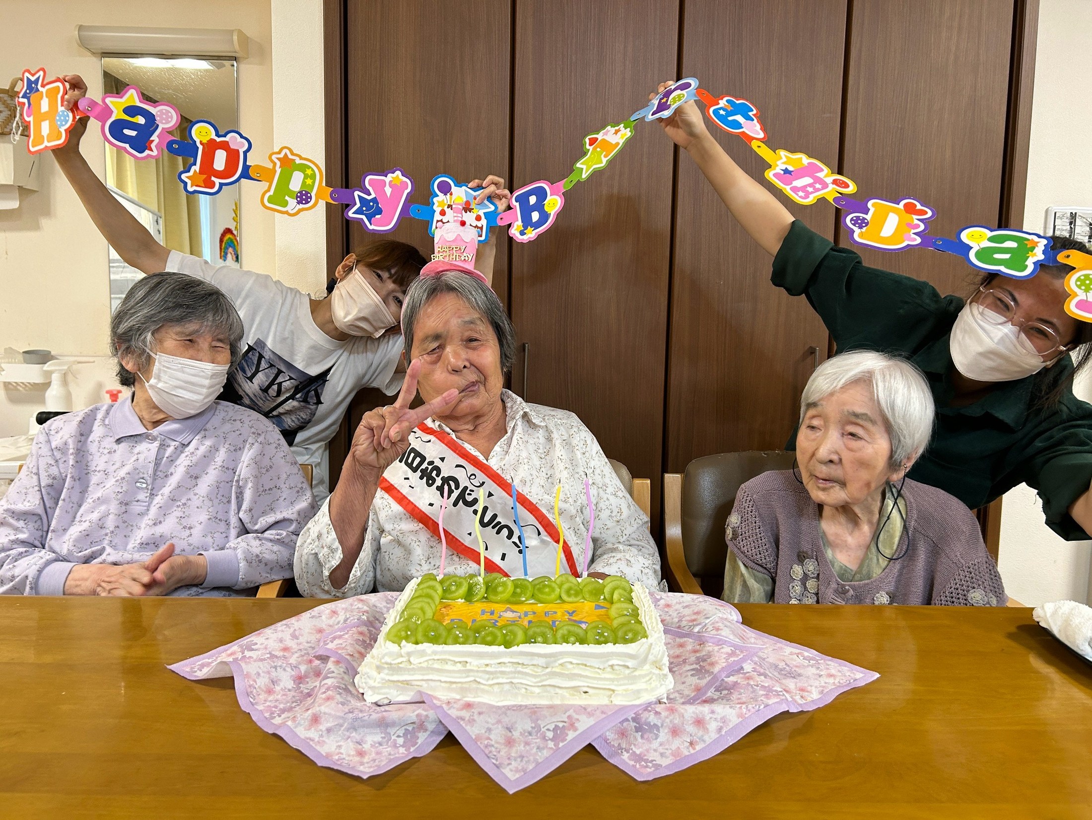 誕生日のたすきをかけた利用者さんを皆さんと職員がお祝いする様子