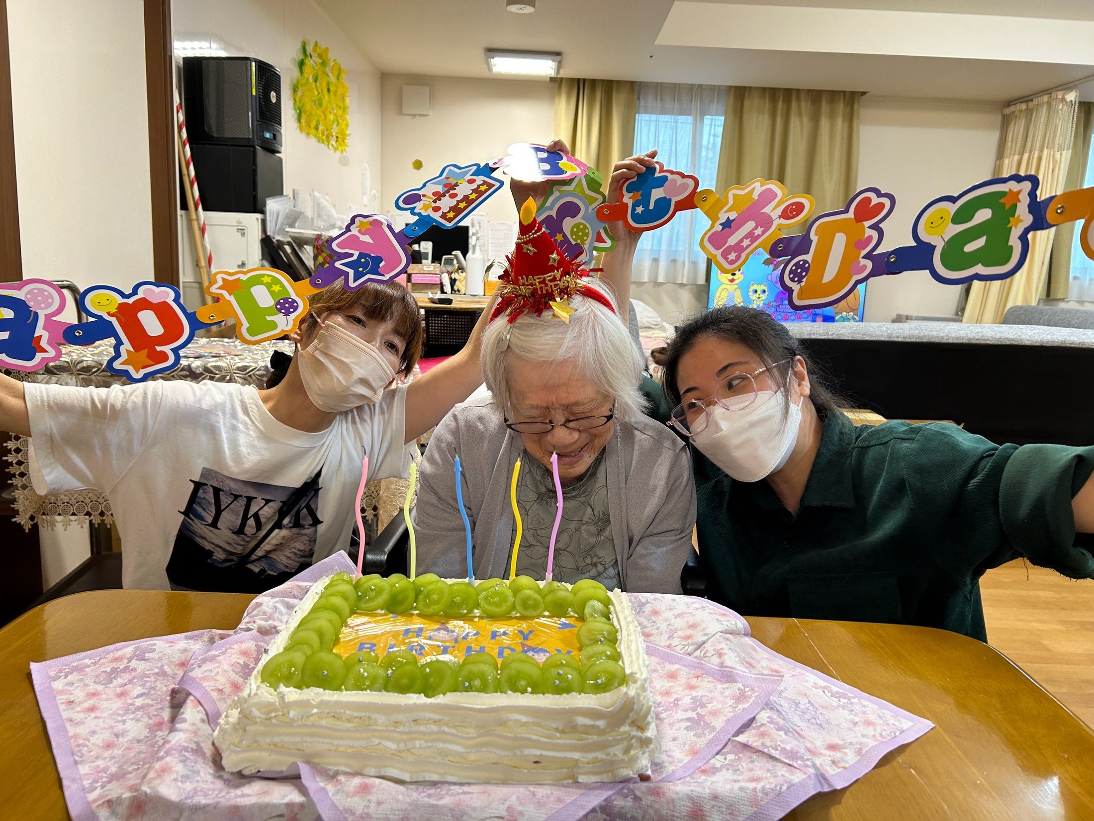 赤い誕生日の帽子をかぶった利用者さんと職員がケーキを囲む様子