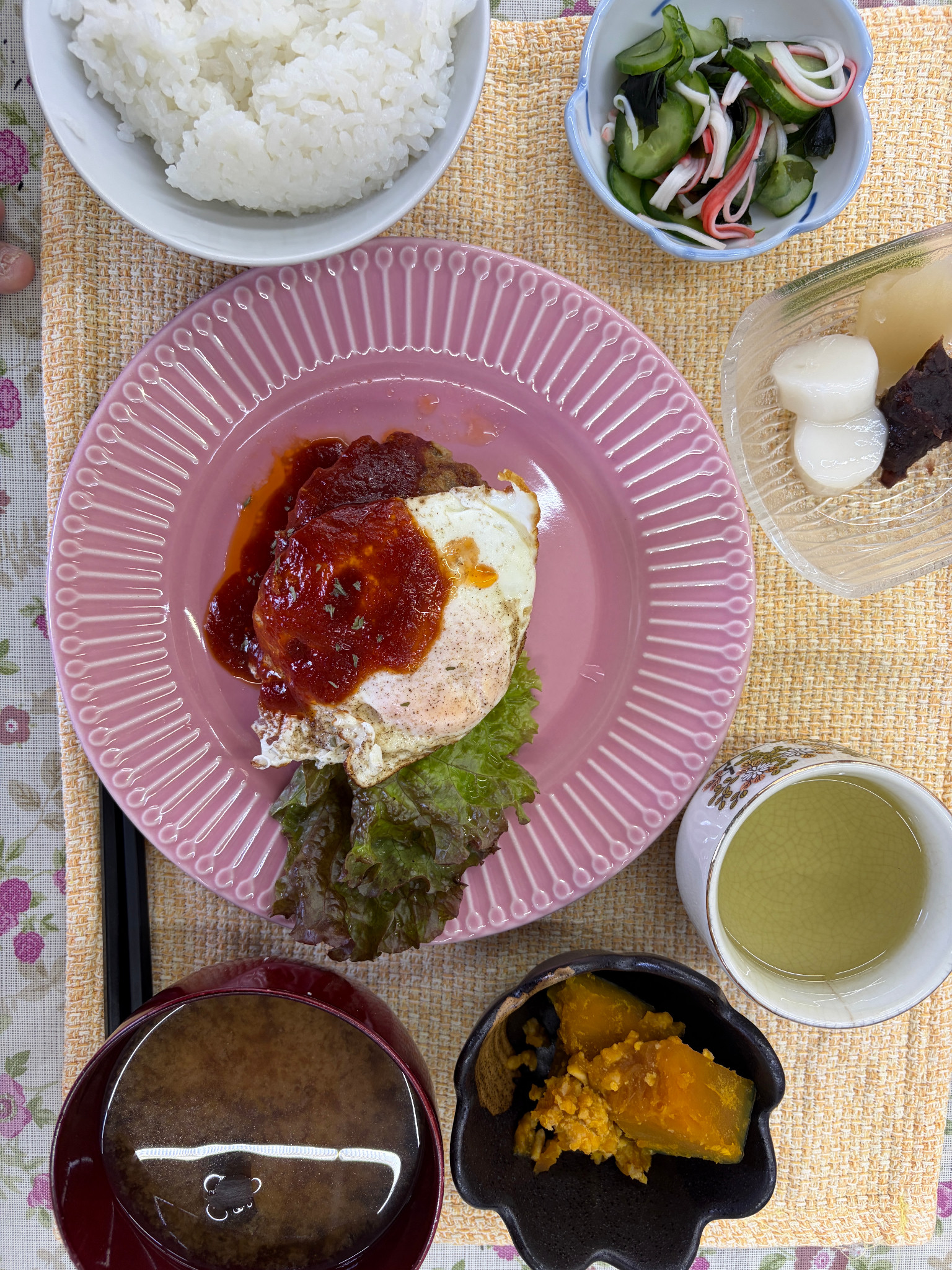 目玉焼きをのせたハンバーグ、ごはん、小鉢、汁もの、甘味が並ぶ食事