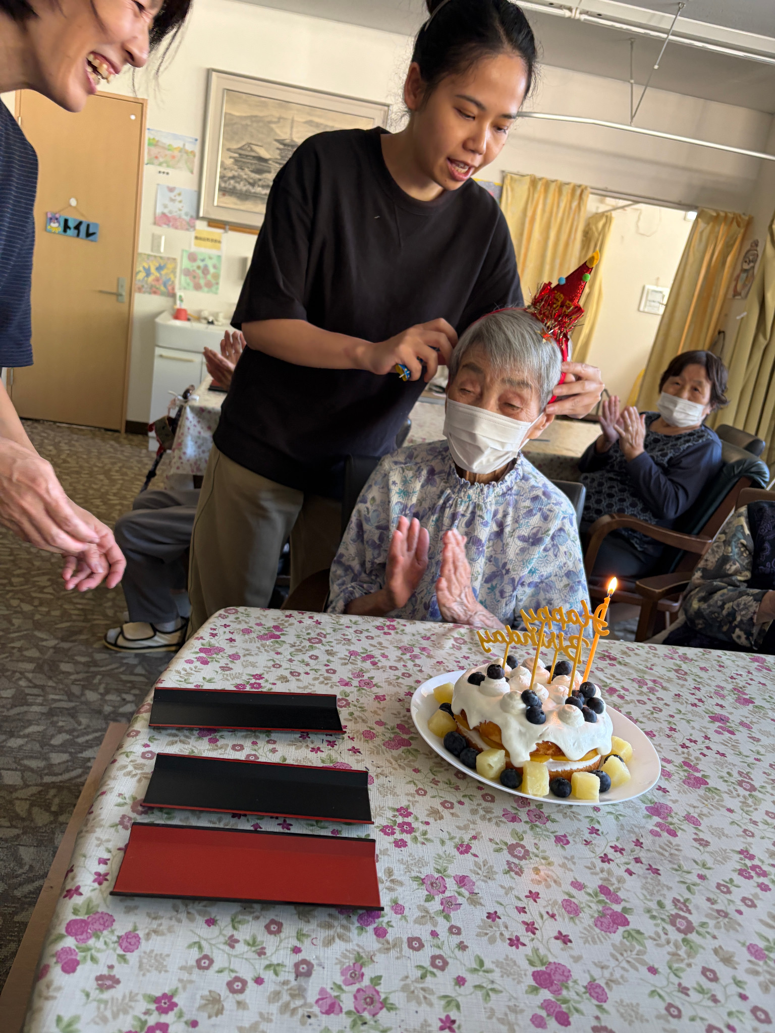 ろうそくの灯る誕生日ケーキを囲み、お祝いの拍手をするS様と皆さん